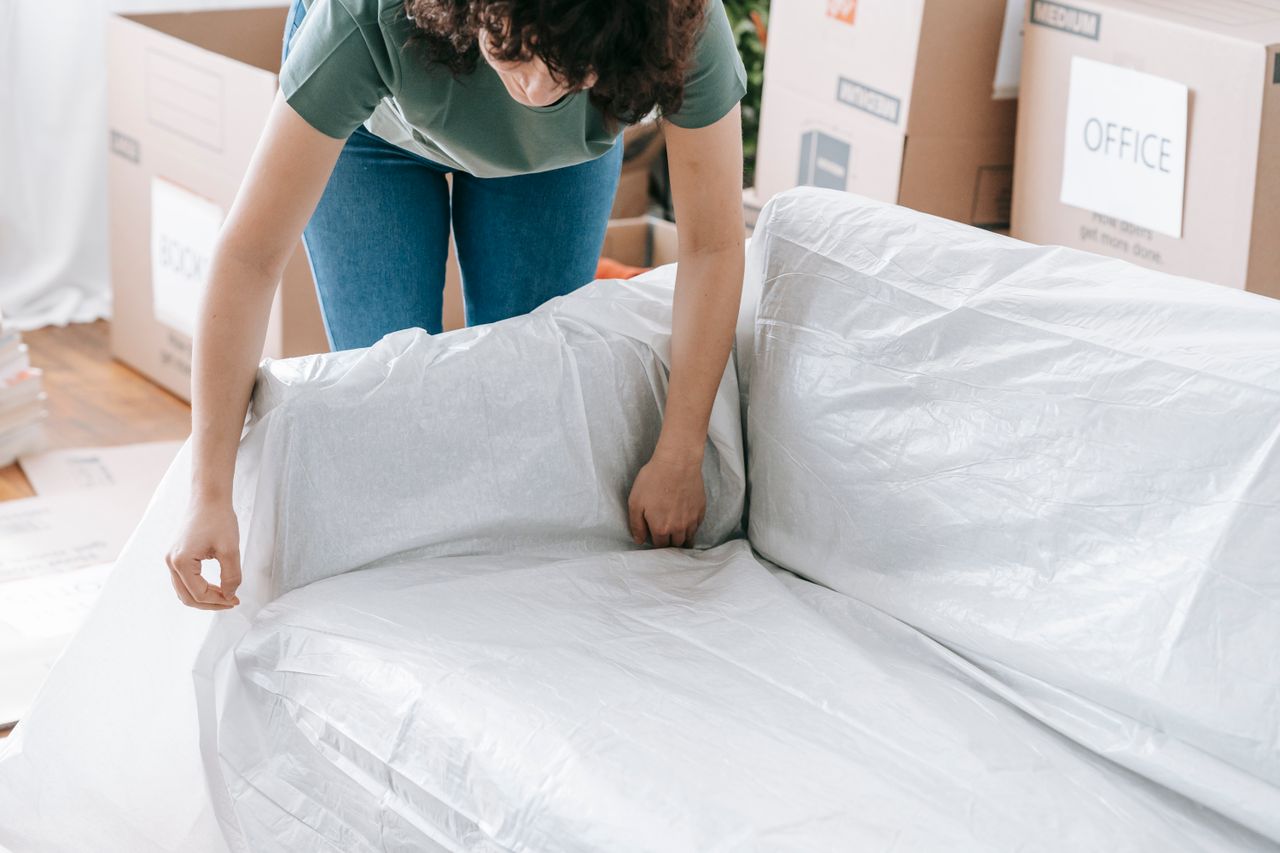services-02 A woman carefully wraps a sofa in protective material while packing boxes in a bright room.